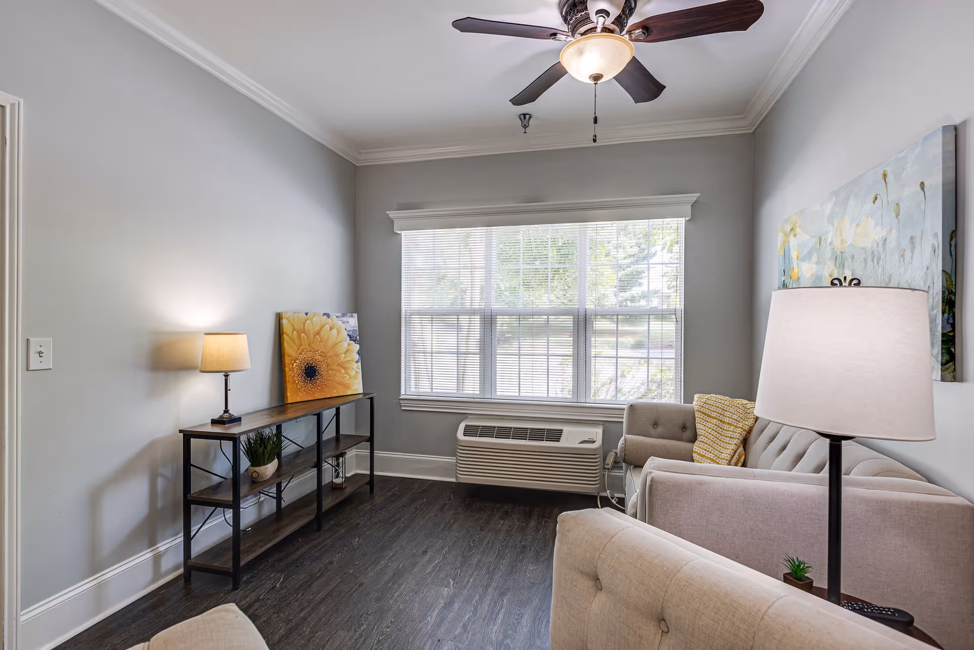 A cozy living room with light gray walls, dark wood flooring, and a large window with blinds. The room features a beige tufted sofa with a yellow patterned pillow, a matching armchair, a floor lamp, a ceiling fan with light, and a narrow console table with a table lamp, a potted plant, and a large yellow flower artwork leaning against the wall.