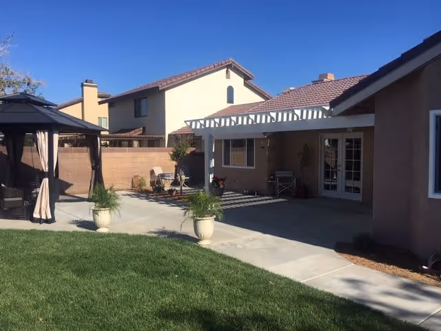 Outdoor patio area of a residential facility with a covered pergola attached to the building, a gazebo with curtains on the left, potted plants along the concrete walkway, and a grassy lawn in the foreground. Neighboring houses with tiled roofs are visible in the background.