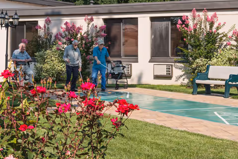 Three elderly men outdoors at a senior living facility playing shuffleboard on a court surrounded by green grass and blooming pink flowers, with a building and benches in the background.