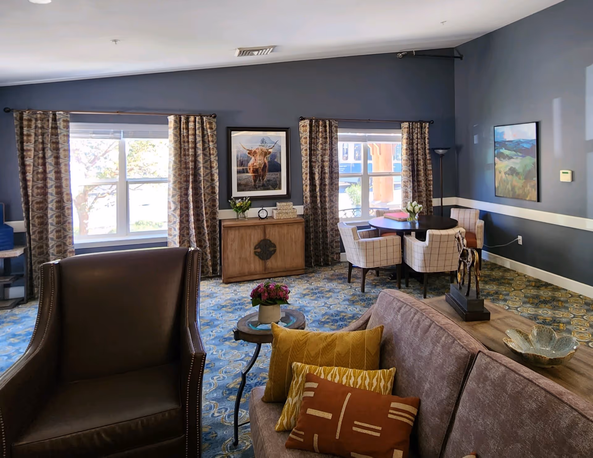 A cozy senior living common area with a brown leather armchair and a sofa adorned with decorative pillows in the foreground. The room features patterned curtains on two windows, a wooden cabinet with a framed picture of a cow above it, and a round table with four upholstered chairs near the windows. The walls are painted gray with white trim, and the floor has a blue and yellow patterned carpet. There are also decorative items including a small sculpture and a flower vase on the tables.