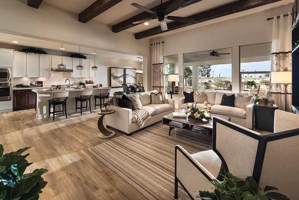 Spacious open-concept living room and kitchen area with wooden ceiling beams, large windows with curtains, two beige sofas with decorative pillows, a coffee table with flowers and candles, a striped area rug, and a kitchen island with bar stools and pendant lights.