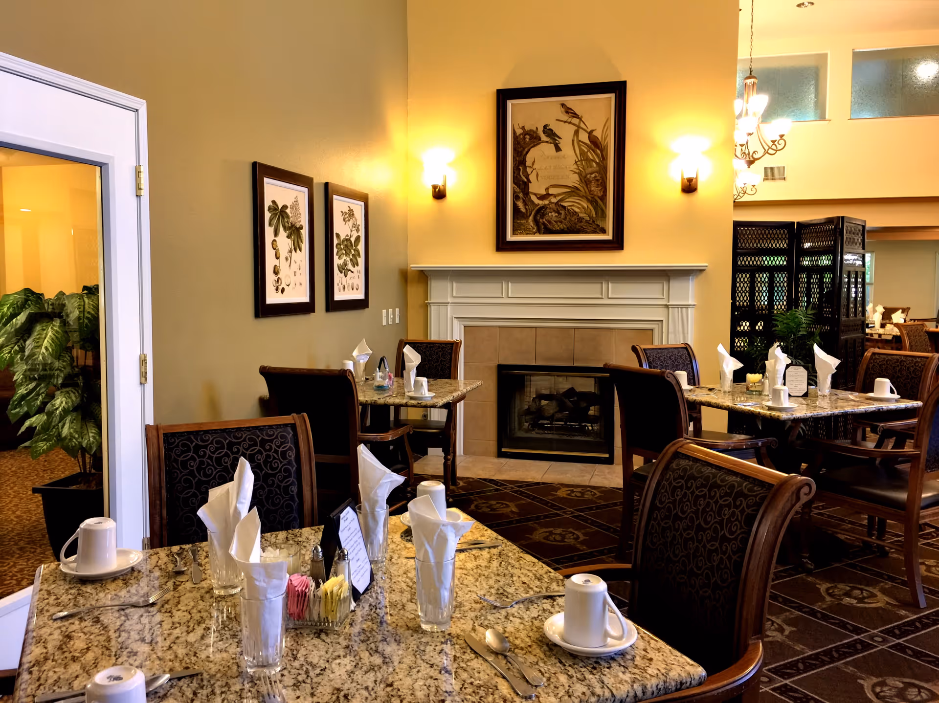 Dining area with granite tables set with cups, glasses with folded napkins, and utensils. The room features upholstered chairs, a fireplace with framed artwork above it, wall sconces providing warm lighting, and decorative plants. There is a glass door on the left and a decorative room divider in the background.