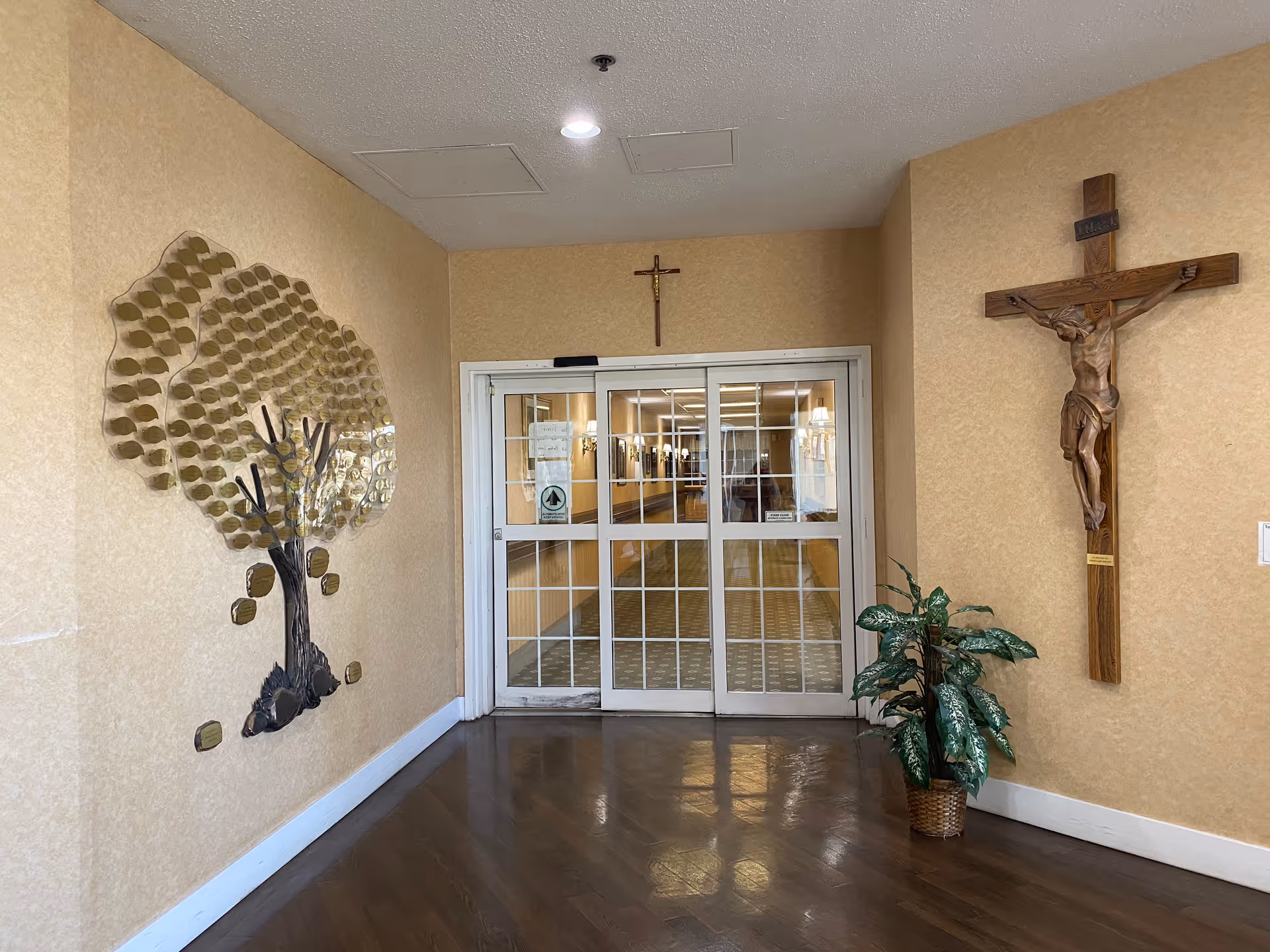 Interior hallway entrance with sliding glass doors. On the left wall, there is a decorative metal tree sculpture with gold leaves. On the right wall, there is a wooden crucifix mounted above a potted plant. The floor is wooden, and the walls are beige.