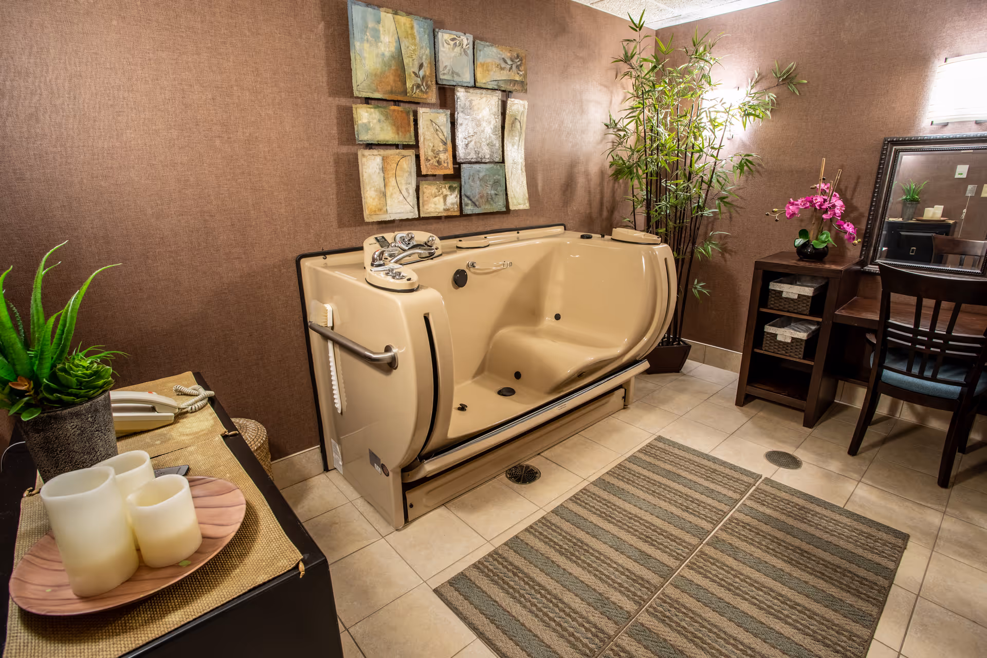 A well-lit bathroom featuring a beige walk-in bathtub with safety handles and a built-in seat. The room has brown textured walls, a decorative multi-panel wall art above the tub, and a tall green plant in the corner. There is a dark wooden vanity with a mirror, a chair, and a pink orchid on top. On the left side, there is a small table with three white candles on a wooden tray and a green potted plant. The floor is tiled with two striped rugs placed in front of the tub and vanity.