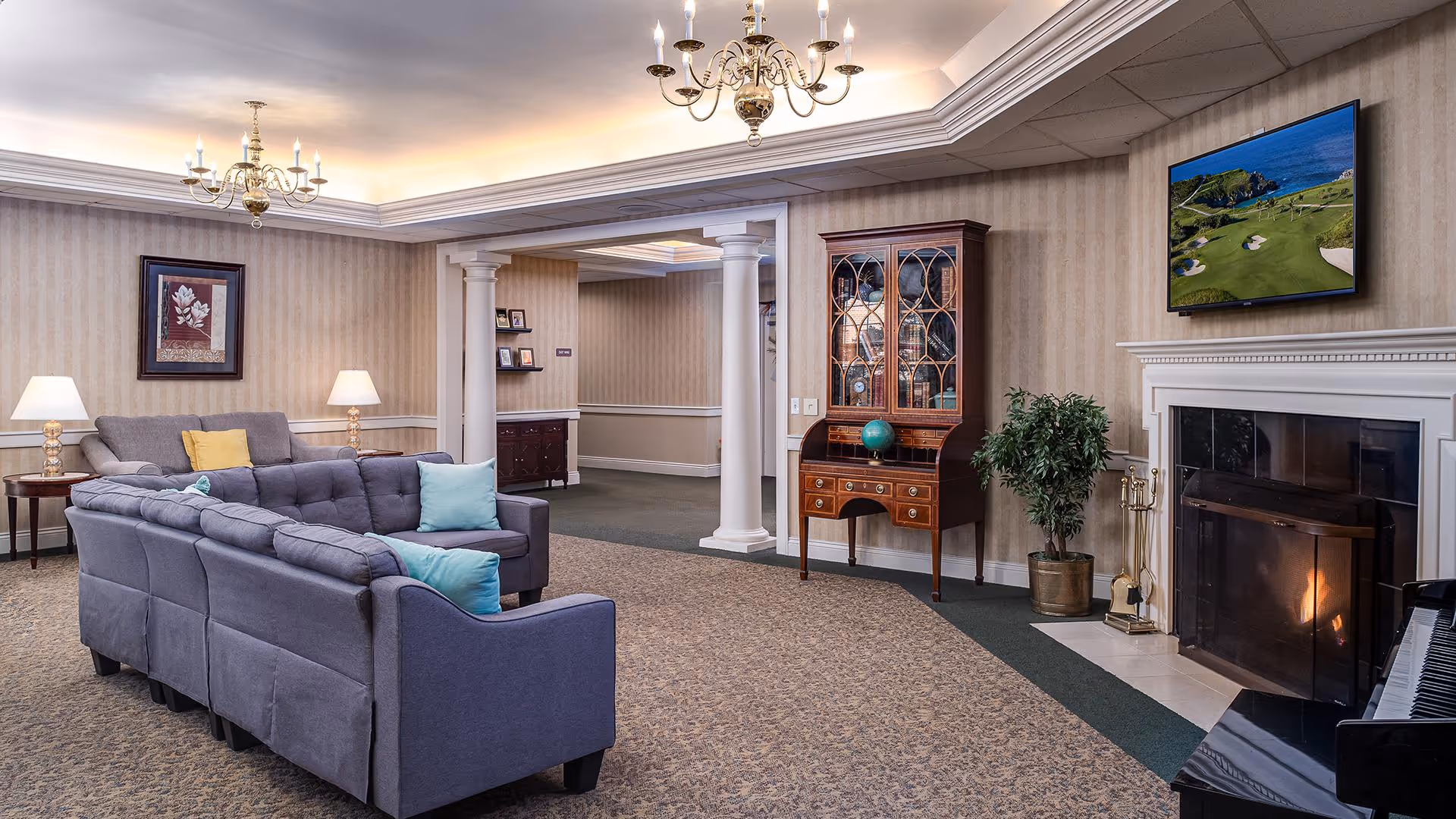 A cozy living room area with a gray sectional sofa adorned with colorful pillows, two side tables with lamps, a framed floral artwork on the wall, a wooden cabinet with glass doors and a globe on top, a fireplace with a fire burning, a flat-screen TV mounted above the fireplace displaying a golf course, a potted plant next to the fireplace, and a piano partially visible on the right side. The room has beige wallpaper, carpeted flooring, and two chandeliers hanging from the ceiling.