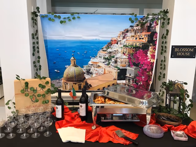 A buffet setup with wine bottles, empty wine glasses, napkins, and a chafing dish with food on a table covered with a black and red cloth. Behind the table is a large scenic backdrop of a coastal town with colorful buildings on a hillside overlooking the sea. Green leafy vines decorate the backdrop and surrounding area. A sign on the wall reads 'Blossom House'.