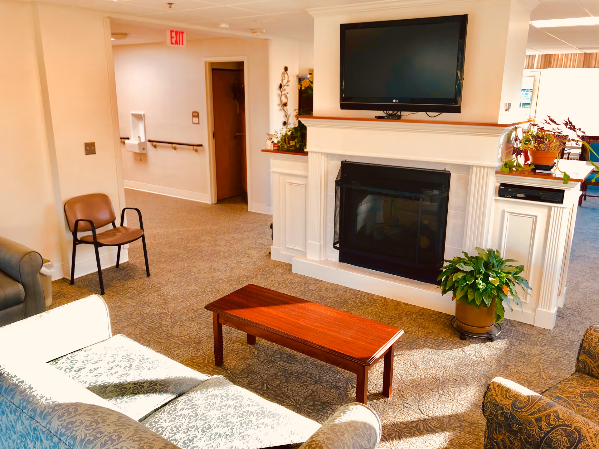 A cozy senior living community lounge area with a patterned carpet, a wooden coffee table, a white fireplace with a black screen, and a flat-screen TV mounted above it. There are several chairs and sofas arranged around the coffee table, potted plants on and near the fireplace, and a hallway with an exit sign in the background.