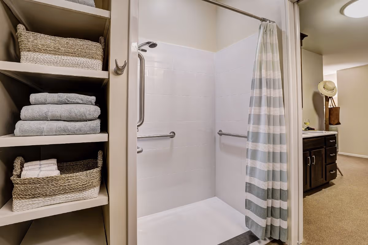 A bathroom shower area with white tiled walls, equipped with metal grab bars and a striped shower curtain. To the left, there are shelves holding folded towels and woven baskets. In the background, part of a room with a cabinet, a hat on a stand, and a glass of wine on the counter is visible.