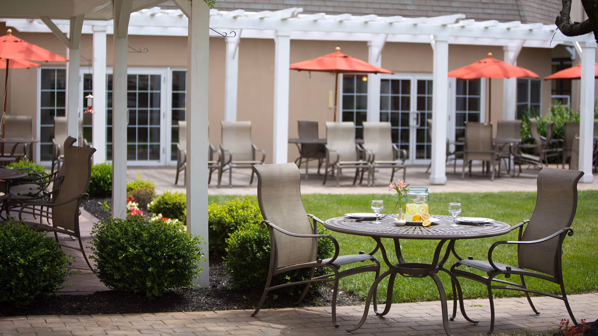 Outdoor patio area with metal table and chairs on a paved surface, a pitcher of lemonade and two glasses on the table, surrounded by green bushes and grass, with beige building and red umbrellas in the background.