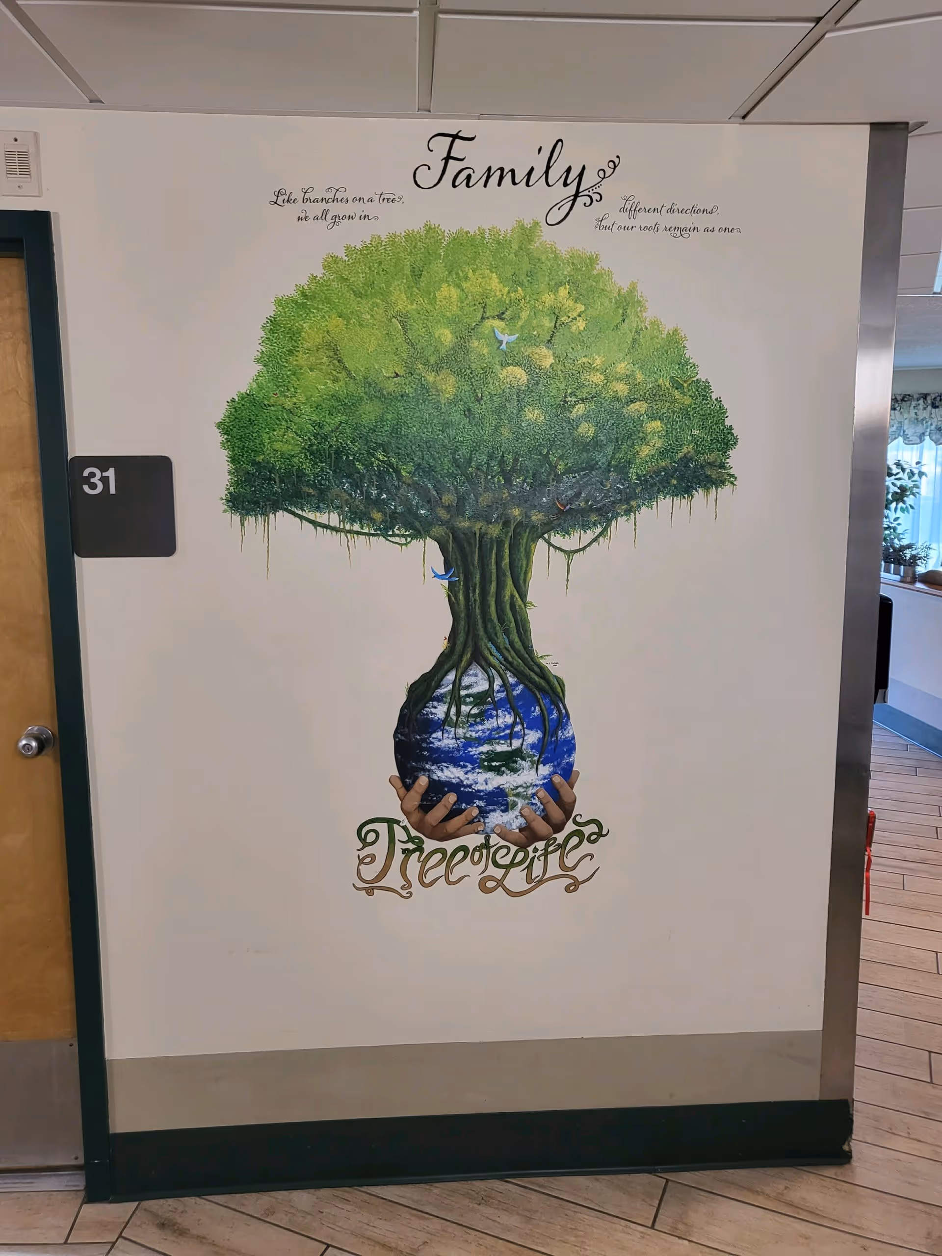A wall mural in a facility hallway depicting a large green tree growing from a globe held by two hands. Above the tree, the word 'Family' is written in cursive, with the phrase 'Like branches on a tree, we all grow in different directions, but our roots remain as one.' Below the globe, the words 'Tree of Life' are written in decorative script. The mural is next to a door labeled with the number 31.