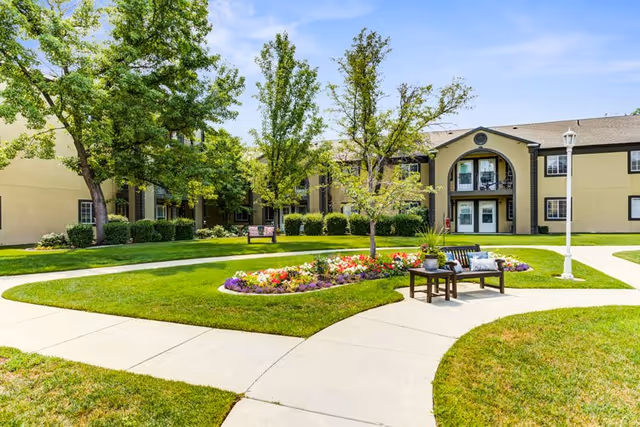 Outdoor courtyard area of Cedarwood at Sandy featuring a well-maintained lawn, a flower bed with colorful flowers, several trees, a bench with cushions, and a paved walkway leading to the building entrance under a clear blue sky.