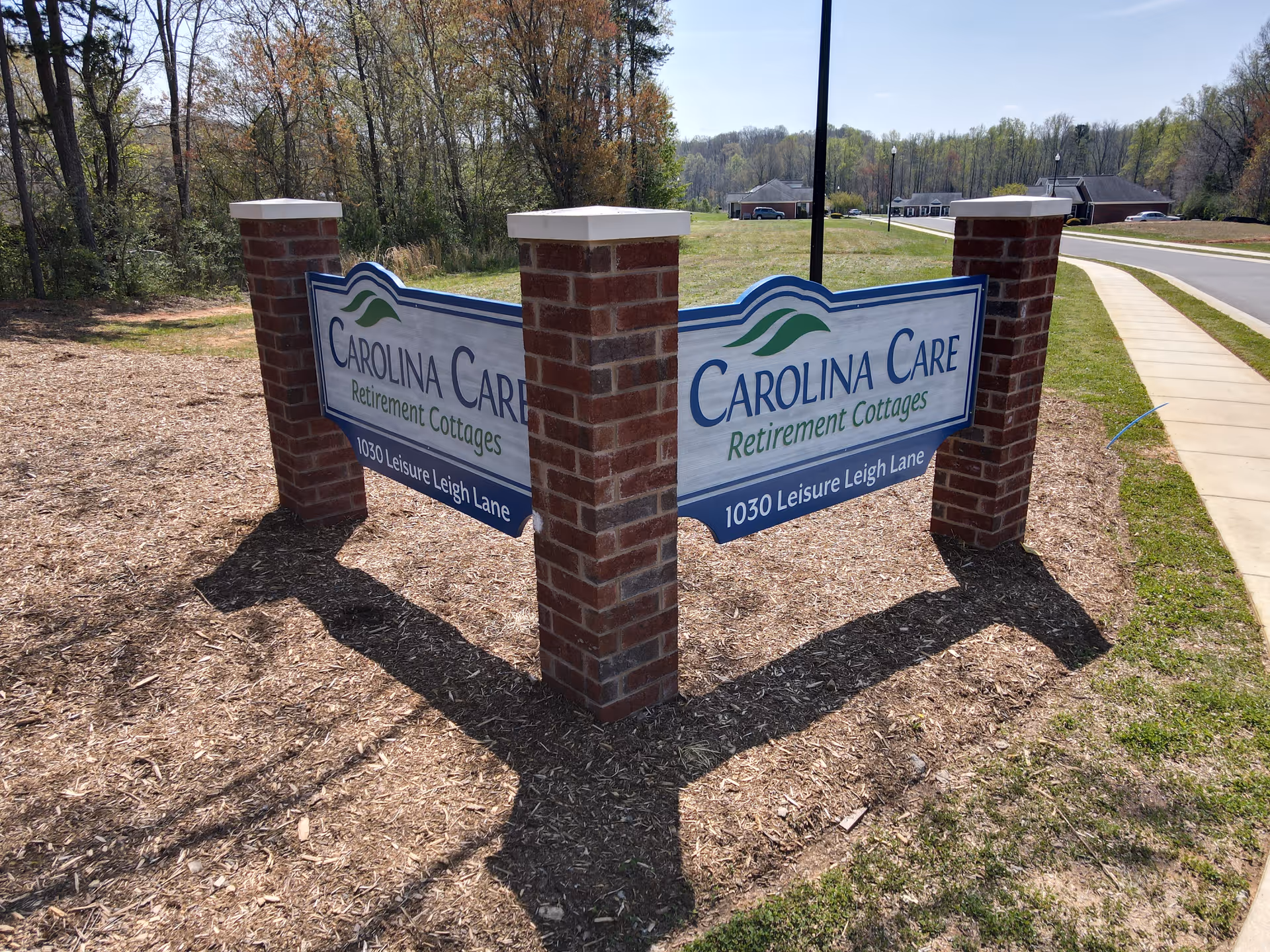 A large outdoor sign for Carolina Care Retirement Cottages mounted on three brick pillars, located near a sidewalk and a road with trees and buildings in the background.