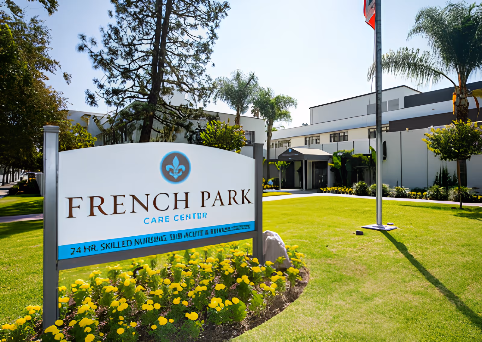 Exterior view of French Park Care Center with a large sign in the foreground surrounded by yellow flowers and a well-maintained green lawn. The building is white with multiple windows, palm trees, and a flagpole with a flag.