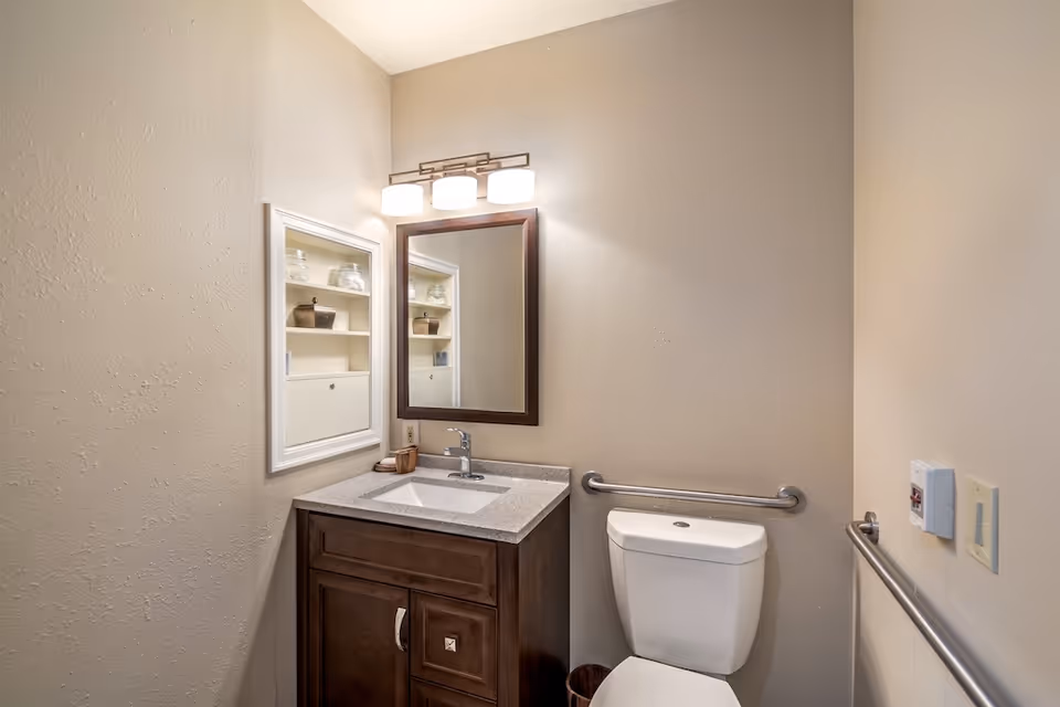 A small bathroom with beige walls featuring a white toilet with a metal grab bar on the wall behind it and another grab bar on the adjacent wall. There is a dark wood vanity with a gray countertop and a built-in sink. Above the sink is a rectangular mirror with a dark wood frame and a three-light fixture mounted above it. To the left of the mirror is a recessed medicine cabinet with shelves holding glass jars and small baskets.