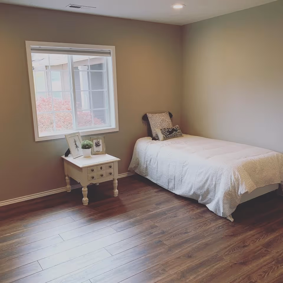 A simple bedroom with a single bed, a small white bedside table, a window, and hardwood floors.