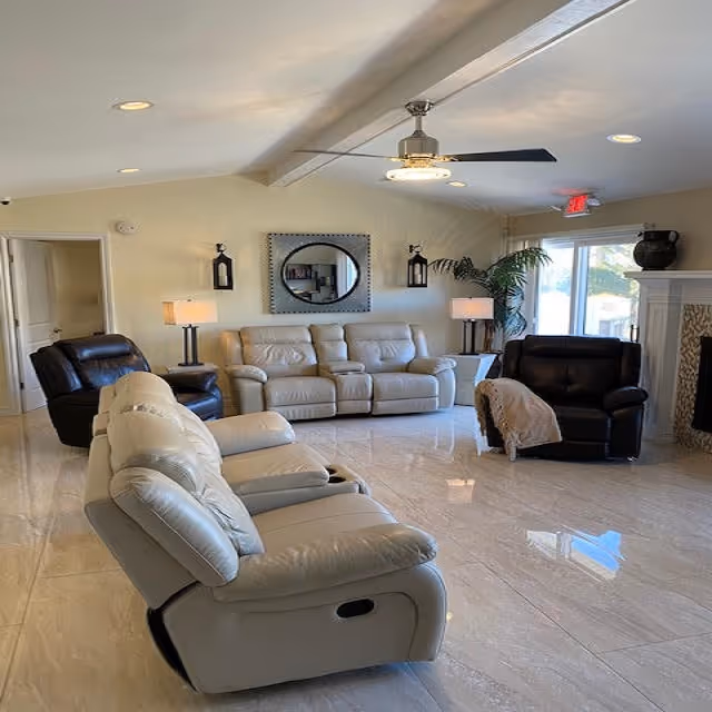 A spacious living room with cream-colored leather recliners and sofas arranged around the room. There are two black leather armchairs, one with a beige throw blanket. The room features a ceiling fan with a light, two table lamps, a large round mirror on the wall, and a potted plant near a sliding glass door. The floor is shiny and tiled, and there is a fireplace on the right side.