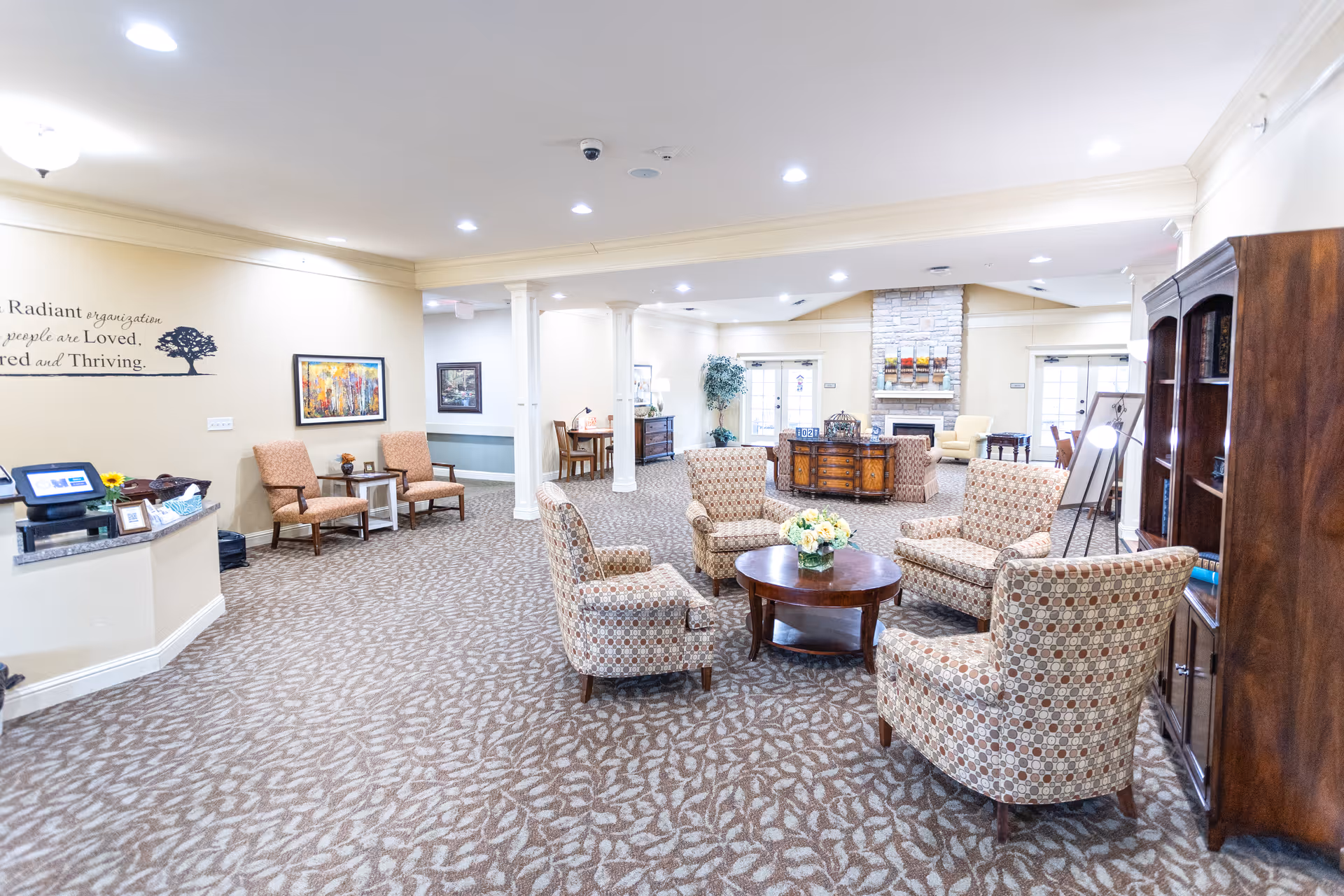A spacious and well-lit senior living facility common area with patterned carpet flooring, several upholstered armchairs arranged around a wooden coffee table with a flower arrangement, a stone fireplace in the background, wooden bookshelves, framed artwork on the walls, and a reception desk on the left side.