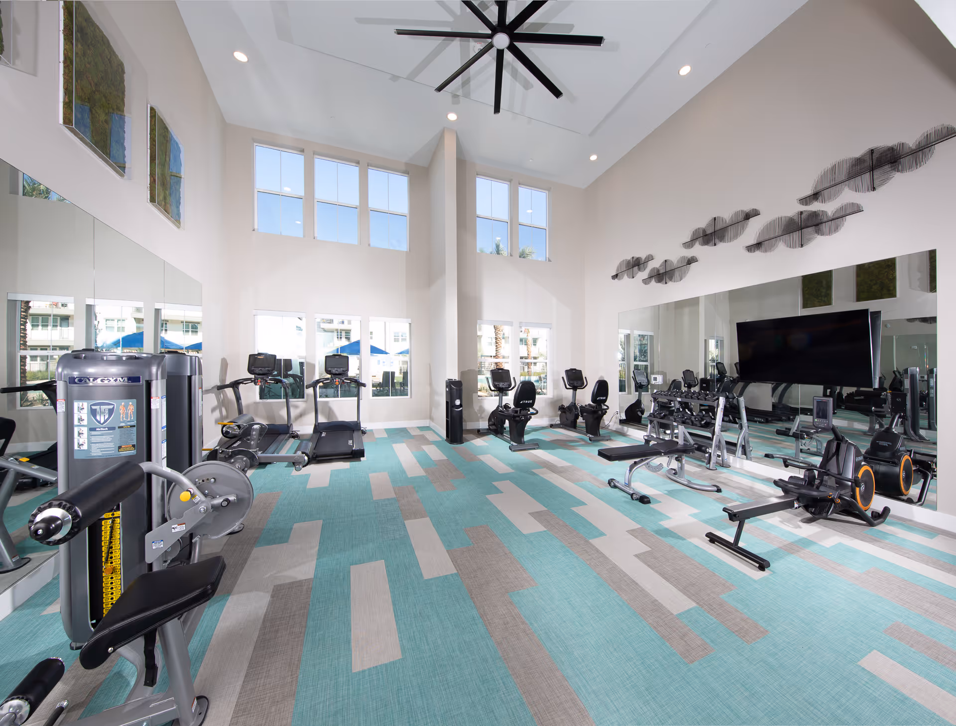 Bright fitness center with exercise machines, treadmills, rowing machines, and large mirrors under tall windows.