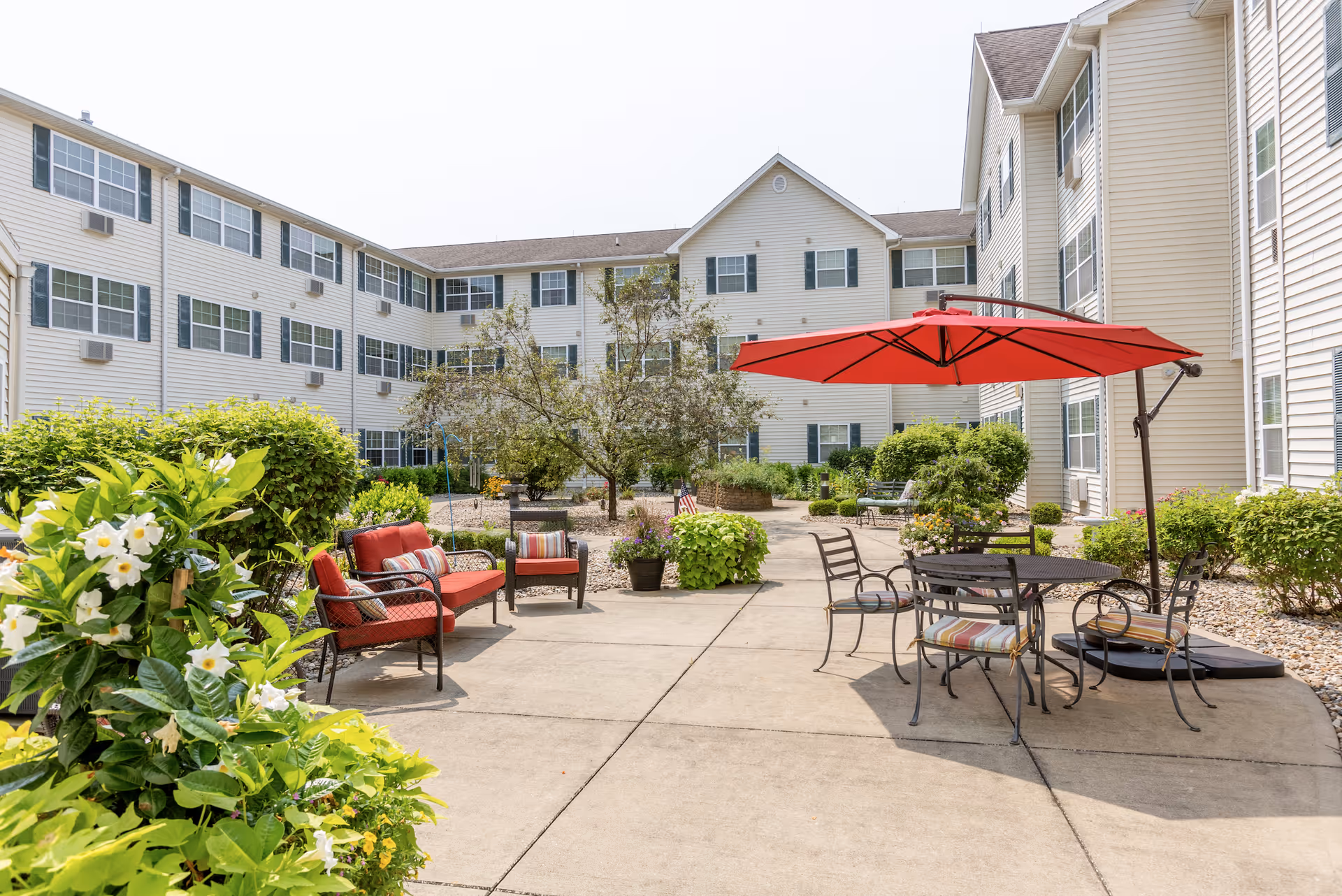Outdoor courtyard area of a senior living facility with patio furniture including red cushioned chairs, a round table with striped cushioned chairs, and a large red umbrella. The courtyard is surrounded by a three-story beige building with multiple windows and green shrubs and trees providing greenery.