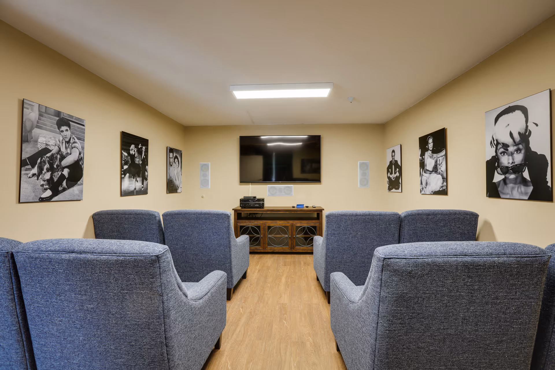 A cozy common room with six blue upholstered armchairs arranged in two rows facing a large flat-screen TV mounted on a beige wall. Below the TV is a wooden cabinet with audio equipment. The walls are decorated with black and white portraits of various people. The room has wood flooring and a rectangular ceiling light.