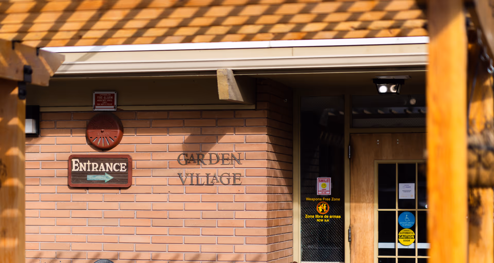 Entrance to Garden Village facility showing a brick wall with a sign that reads 'ENTRANCE' with an arrow pointing right, and the words 'GARDEN VILLAGE' mounted on the wall. There is a wooden door with glass panels and several warning and informational stickers, including a 'Weapons Free Zone' sign. The entrance is partially shaded by a wooden pergola structure.
