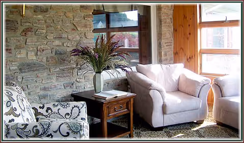 A cozy living room area with a stone wall, a window, two upholstered armchairs, a wooden side table with a vase of flowers and a book, and a patterned carpet.
