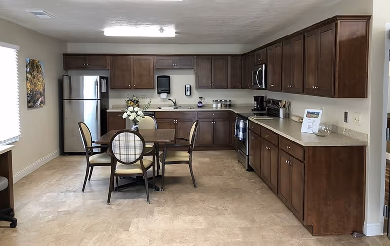 A kitchen area with wooden cabinets and beige countertops. There is a stainless steel refrigerator on the left, a stove with a microwave above it on the right, and a sink in the middle. A small dining table with four chairs is placed in the center of the room, with a vase of white flowers on the table. The floor is tiled, and there is a window with blinds on the left wall.