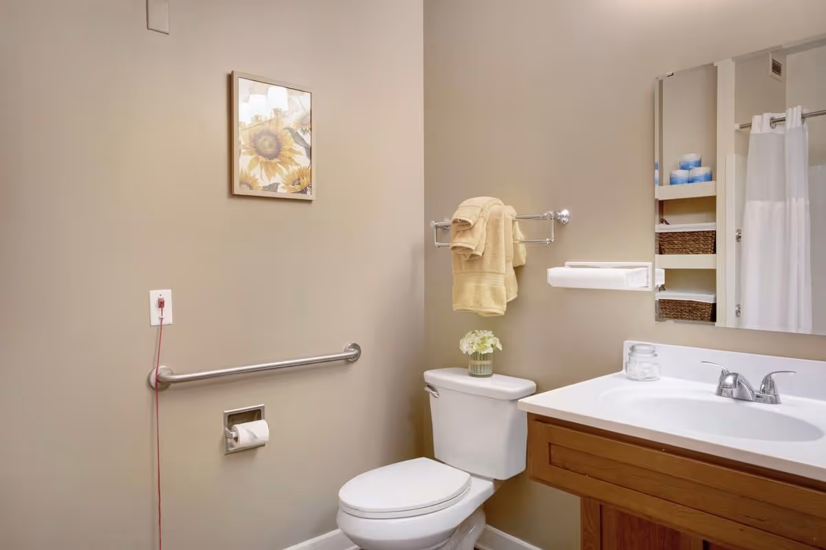 A clean and well-lit bathroom with beige walls featuring a white toilet with a small flower vase on the tank, a towel rack with folded yellow towels, a white sink with a wooden cabinet, a large mirror, and a shower curtain visible in the reflection. There is also a safety grab bar on the wall and a framed sunflower picture hanging above it.