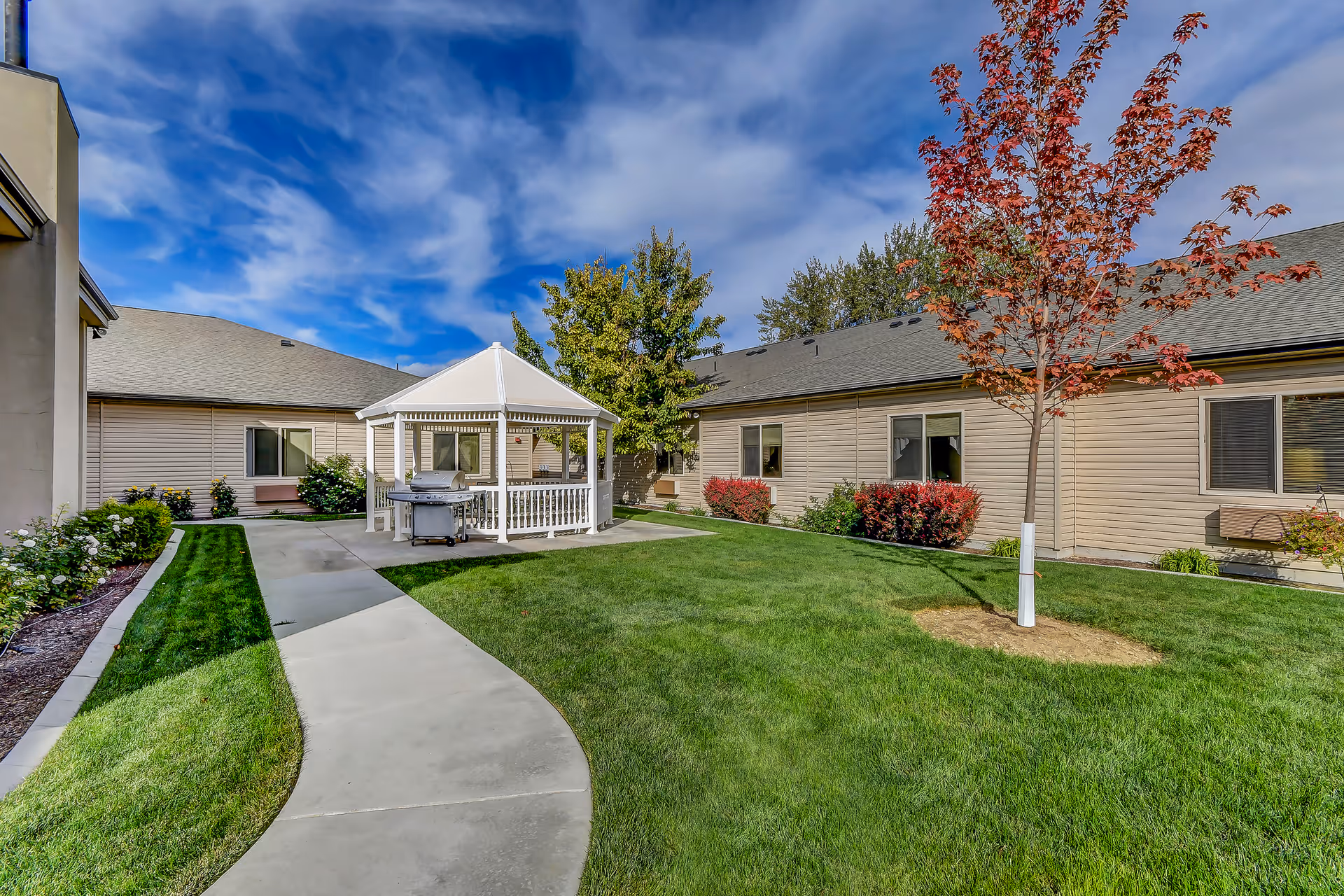 Outdoor courtyard area of Grace Assisted Living - Nampa featuring a curved concrete walkway, green lawn, a white gazebo with a grill inside, surrounded by beige single-story buildings, trees, and shrubs under a partly cloudy blue sky.