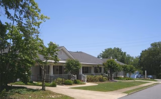 Exterior view of a single-story senior living facility building with a gray roof, white porch railings, and several small trees and bushes in front. The sky is clear and blue, and a paved walkway leads to the entrance.