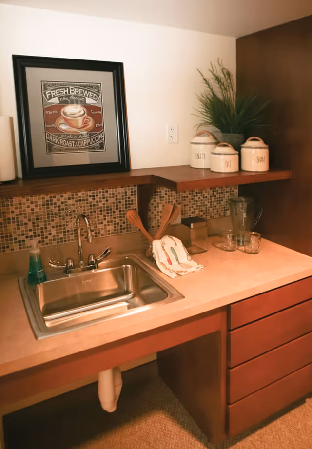 A small kitchen area with a stainless steel sink, a soap dispenser, wooden utensils in a holder, a dish towel, a glass pitcher, and two glass cups on the countertop. Above the counter is a mosaic tile backsplash, a framed coffee-themed picture, three labeled ceramic canisters, and a potted plant.