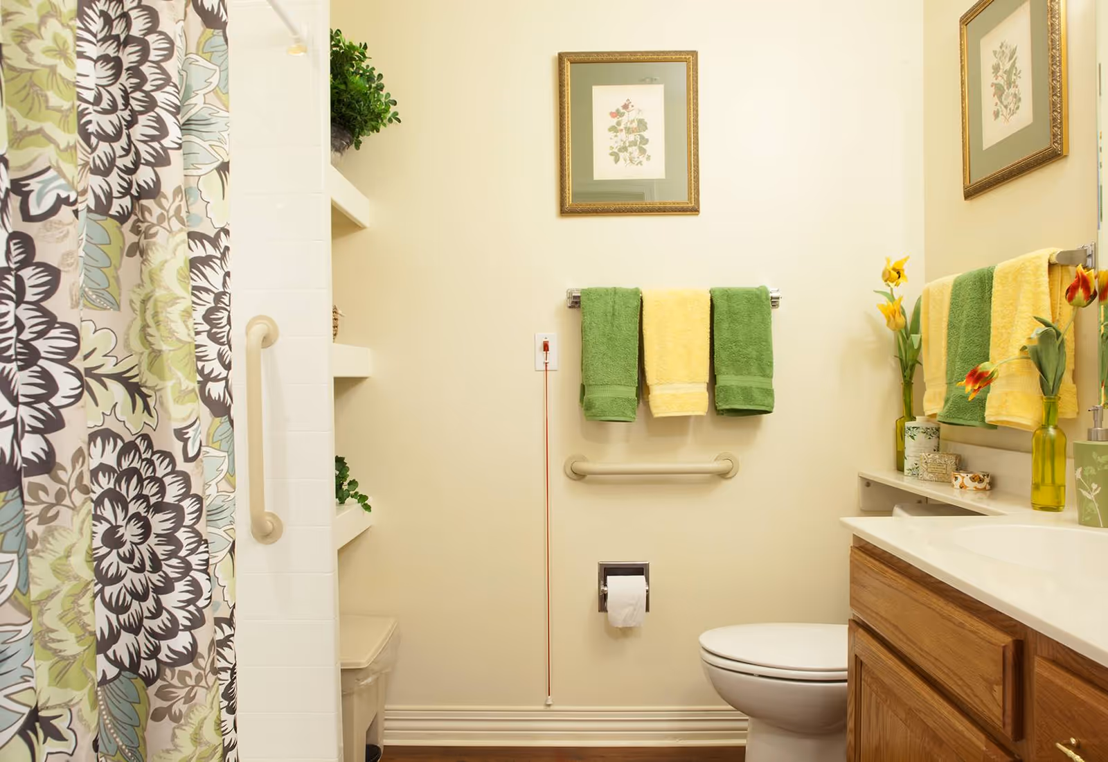A clean and well-lit bathroom featuring a toilet, a wooden vanity with a white countertop, and a shower with a floral patterned curtain. The wall has two towel racks holding green and yellow towels, a grab bar, a framed botanical print, and a red emergency pull cord. There are decorative plants and flowers placed on shelves and the countertop.