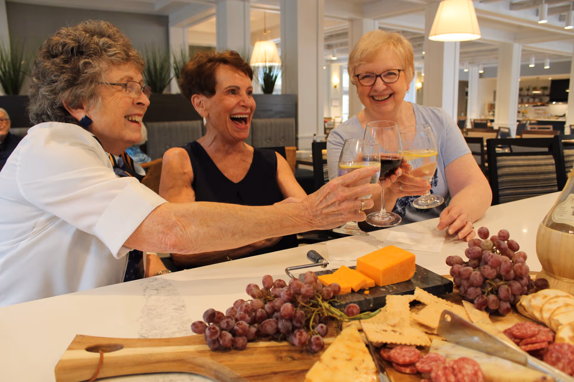 Three older women sit in a dining area, smiling and toasting with wine glasses over a spread of cheese, grapes, crackers, and charcuterie.