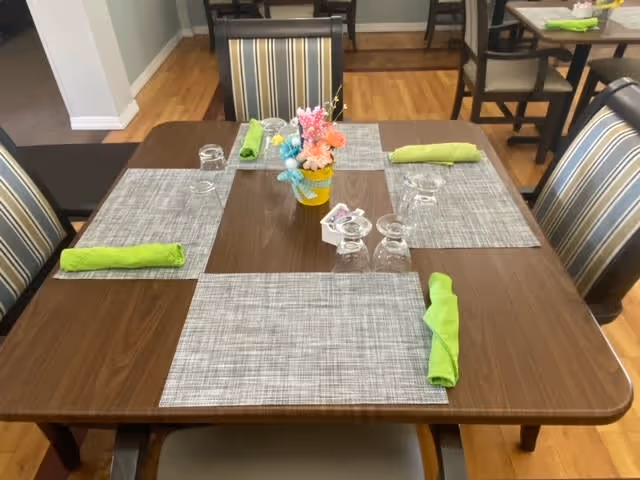 A wooden dining table set for four with gray placemats, green cloth napkins, upside-down drinking glasses, and a small yellow vase with colorful flowers in the center. The chairs around the table have striped upholstery. Additional tables and chairs are visible in the background on a wooden floor.