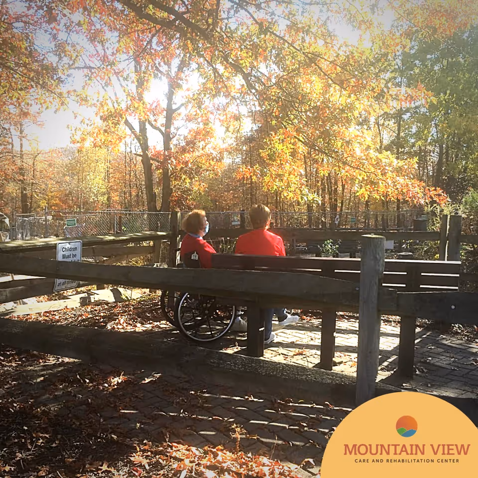 Two elderly individuals sitting on a bench in an outdoor garden area surrounded by autumn trees with colorful leaves. One person is in a wheelchair and both are wearing red jackets. The area is enclosed by wooden fencing and there is a sign that partially reads 'Children Must be...'.
