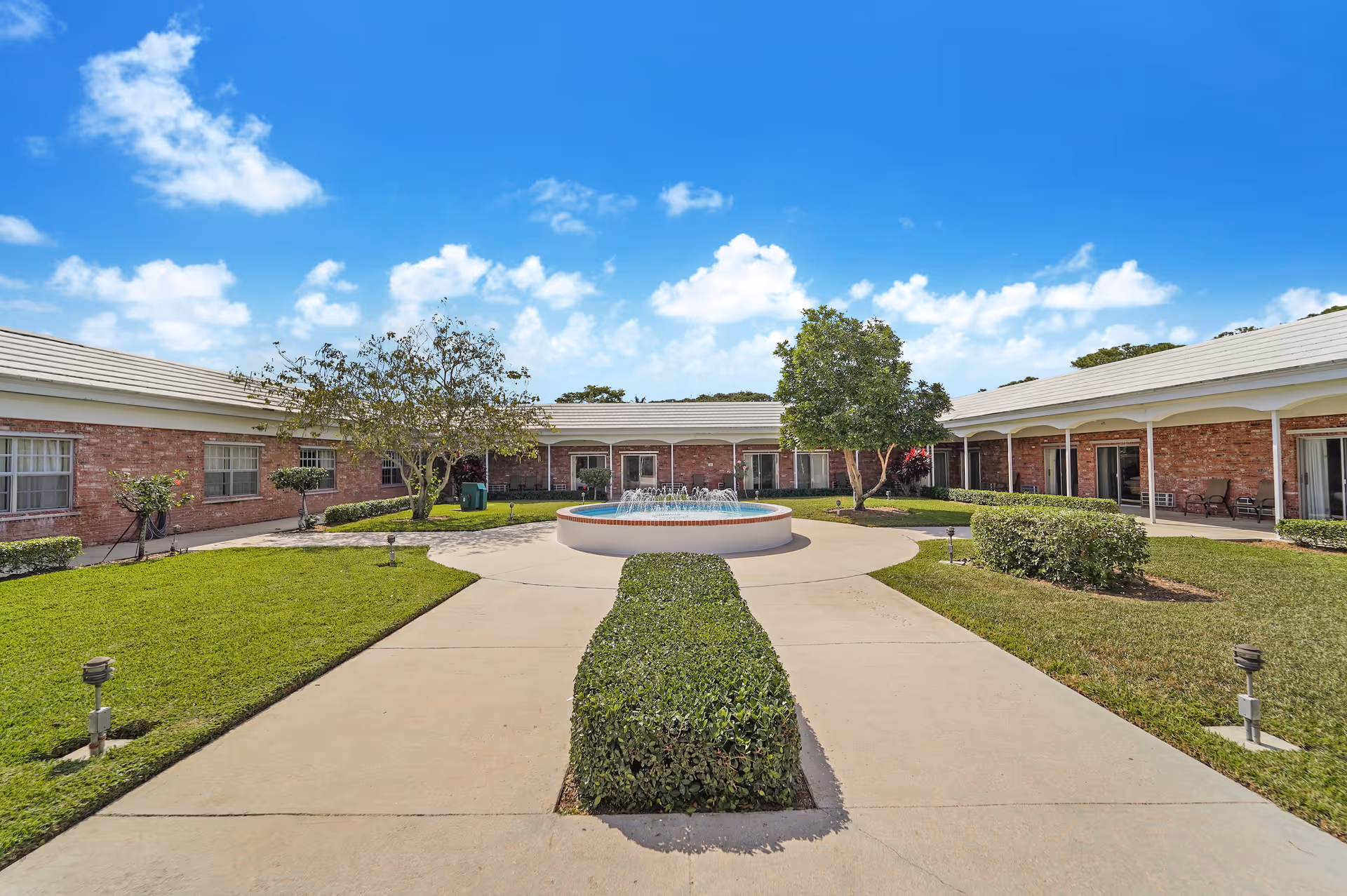 A courtyard area at Manor Pines featuring a central circular water fountain surrounded by a paved walkway and green hedges. The courtyard is enclosed by a single-story brick building with windows and covered walkways. There are a few trees and neatly trimmed grass under a bright blue sky with scattered clouds.