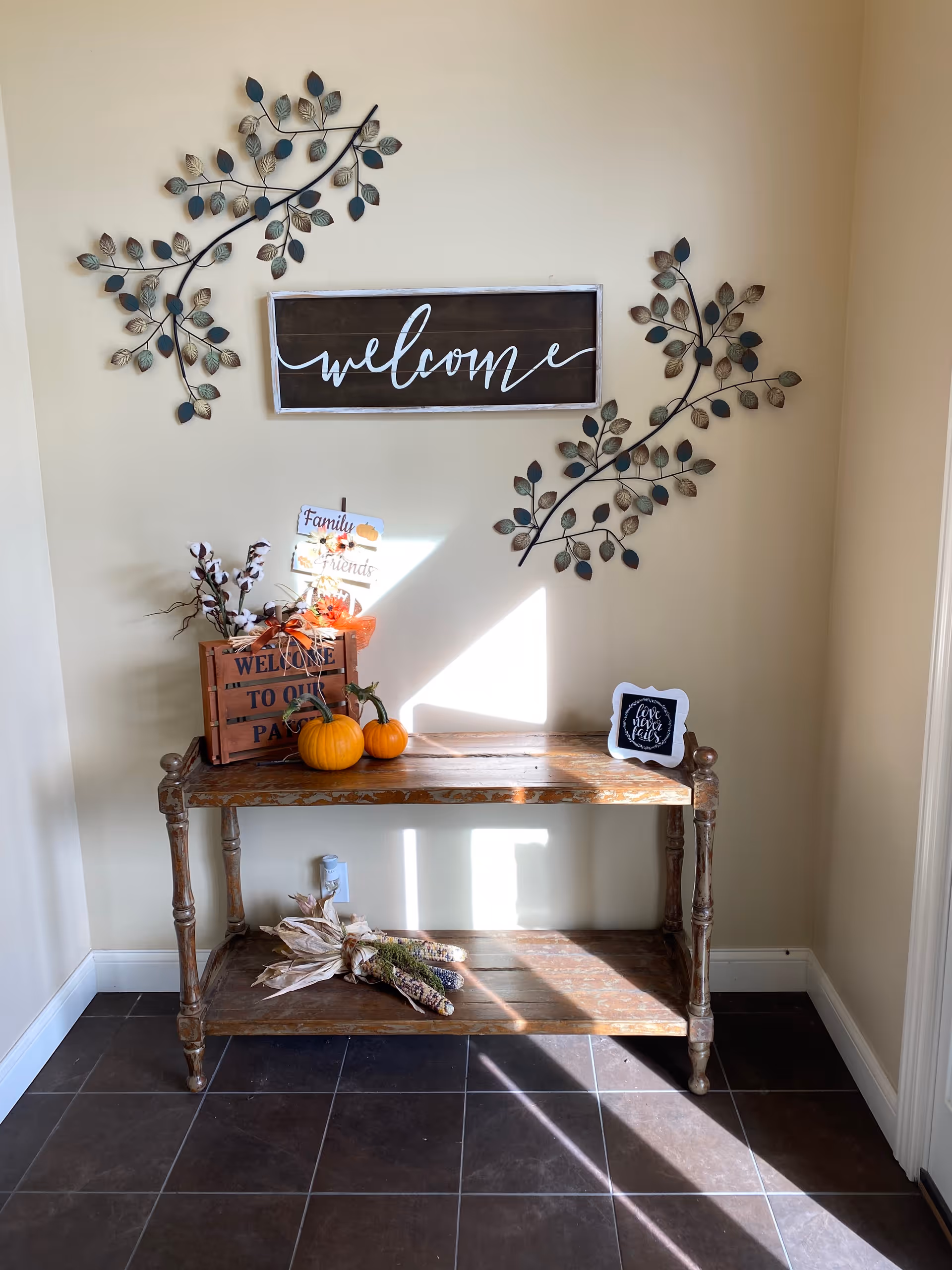 A decorated entry console table beneath a wooden "welcome" sign and metal leaf wall art, with pumpkins and seasonal decorations.