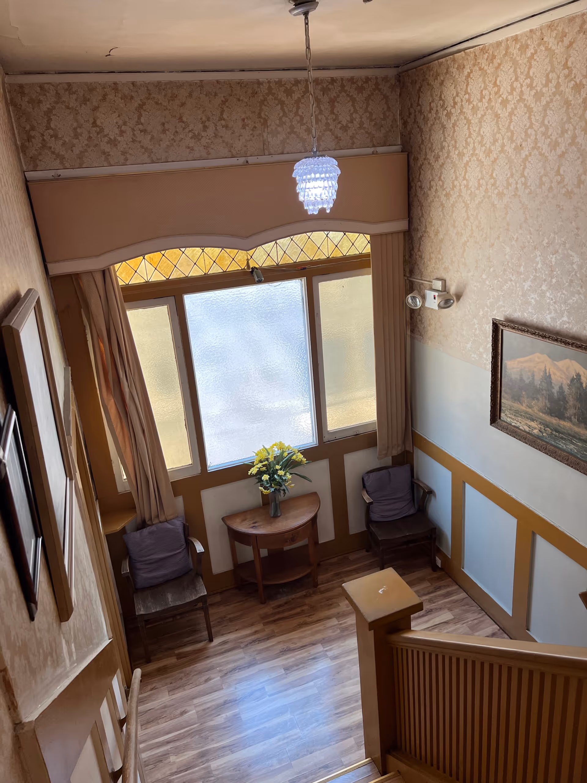 View from the top of a staircase looking down into a small sitting area with two wooden chairs with cushions, a small wooden table with a vase of yellow flowers, and large frosted windows with decorative stained glass at the top. The walls have patterned wallpaper and a framed landscape painting, and a hanging light fixture is visible above.