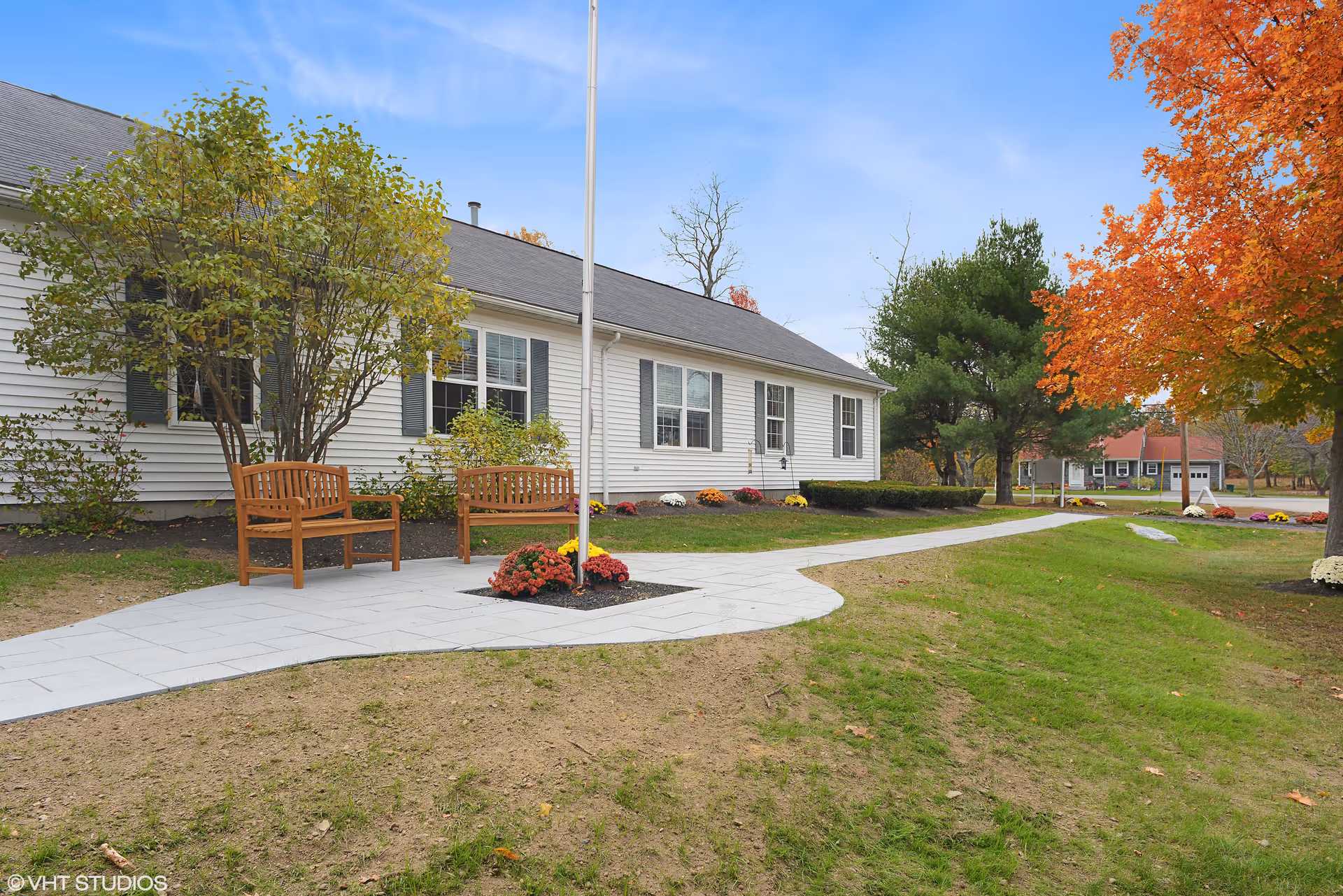 Single-story white building with a paved walkway, wooden benches, a flagpole, and autumn trees on the landscaped front lawn.