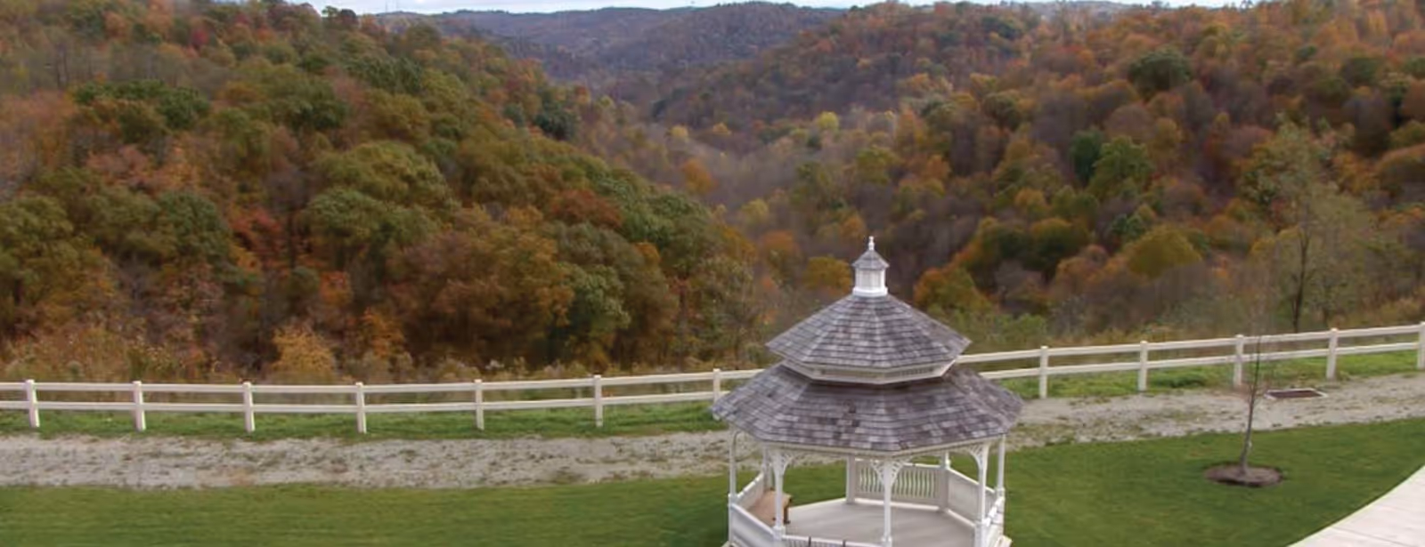 White gazebo on a grassy lawn overlooking a fenced valley with wooded hills showing autumn colors.