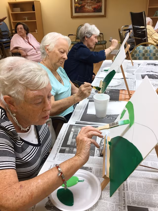 Several elderly women seated at a table in a communal room, engaged in painting on canvases placed on small easels. The table is covered with newspapers, and there are paint palettes and cups of water in front of them. The room has beige walls with a framed picture and stacked chairs in the background.