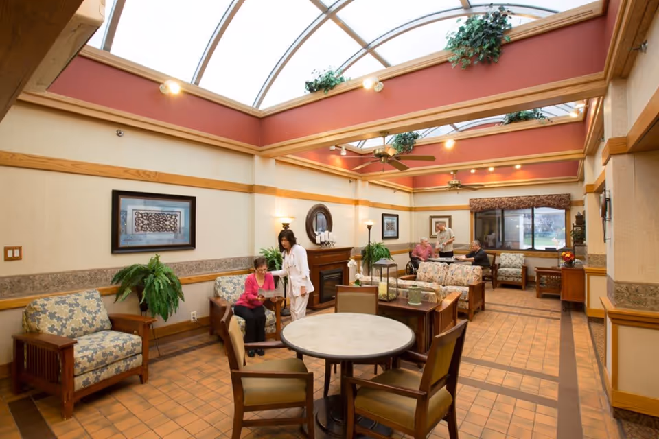A bright and spacious common area in Northgate Health Care Facility with a high glass ceiling, ceiling fans, and warm lighting. The room features comfortable floral-patterned armchairs and sofas arranged around a fireplace and tables. Several people are engaged in conversation, including a caregiver assisting a seated woman and two men talking near the window. The space is decorated with plants, framed artwork, and has a tiled floor.
