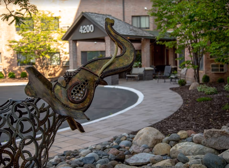 Outdoor view of the entrance to a building with the number 4200 displayed above the doorway. In the foreground, there is a decorative metal sculpture of an elephant's head. The area around the entrance features a paved walkway, landscaping with rocks and plants, and outdoor seating.