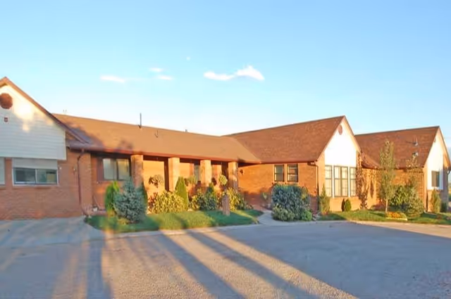 Front exterior of a single-story brick assisted living building with a parking area and landscaped shrubs under a clear blue sky.