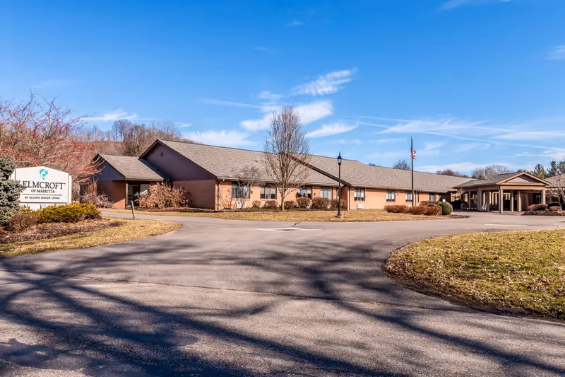Single-story brick assisted living facility with a circular driveway, entrance canopy, flagpole, and a lawn under a blue sky.