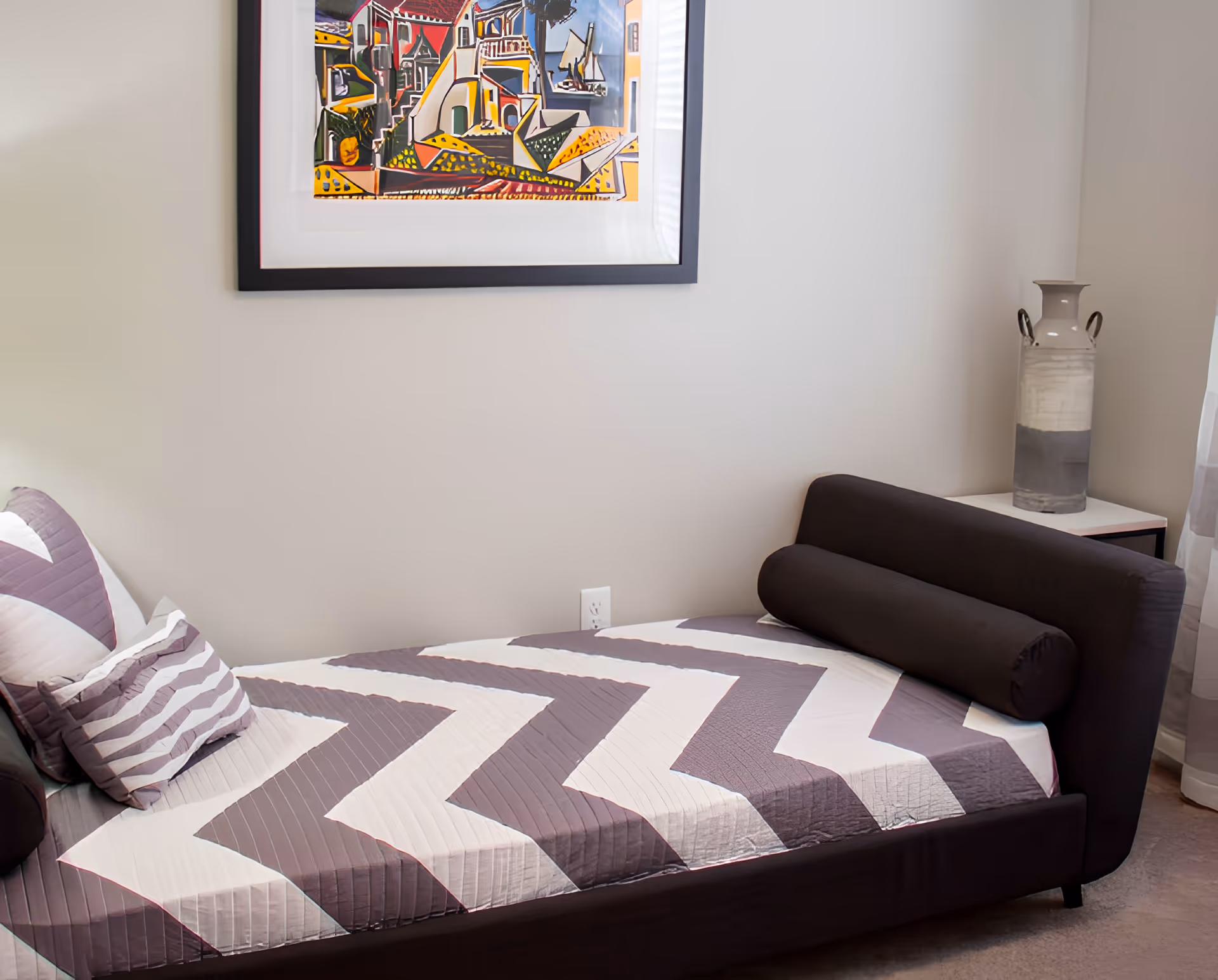 A small bedroom with a single bed featuring a zigzag patterned bedspread in shades of gray and white. The bed has matching pillows and a cylindrical bolster pillow. Above the bed, there is a framed colorful abstract painting. To the right of the bed, there is a small white side table with a tall decorative vase. The room has light-colored walls and carpeted floor.