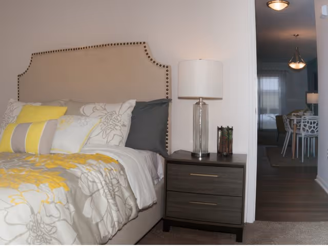 A bedroom with a beige upholstered headboard decorated with nailhead trim, a bed with floral and yellow patterned bedding, and a dark wooden nightstand with two drawers. On the nightstand is a clear glass lamp with a white lampshade and a small decorative item. In the background, a dining area with a table and chairs is visible through an open doorway.