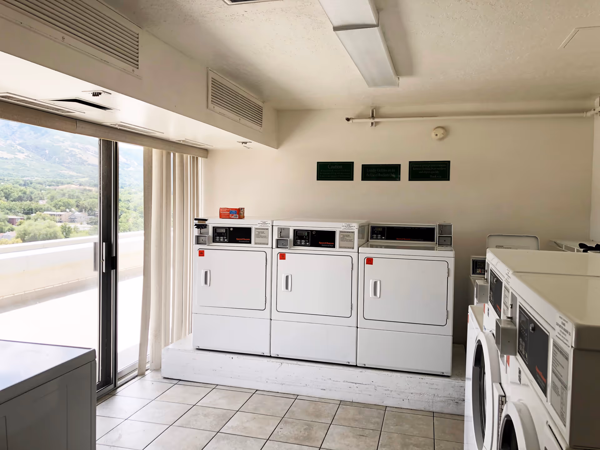 Laundry room with multiple white commercial washing machines and dryers. The room has tiled flooring, a large sliding glass door with vertical blinds, and a view of greenery and mountains outside. There are three green signs on the wall above the machines.