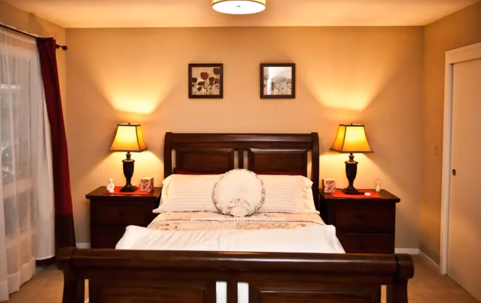 A warmly lit bedroom featuring a wooden bed with white and beige bedding, a round decorative pillow in the center, two matching wooden nightstands each with a lamp emitting soft yellow light, and two framed floral artworks hanging on the beige wall above the bed. There is a window with sheer white curtains and dark red drapes on the left side and a white sliding door on the right.