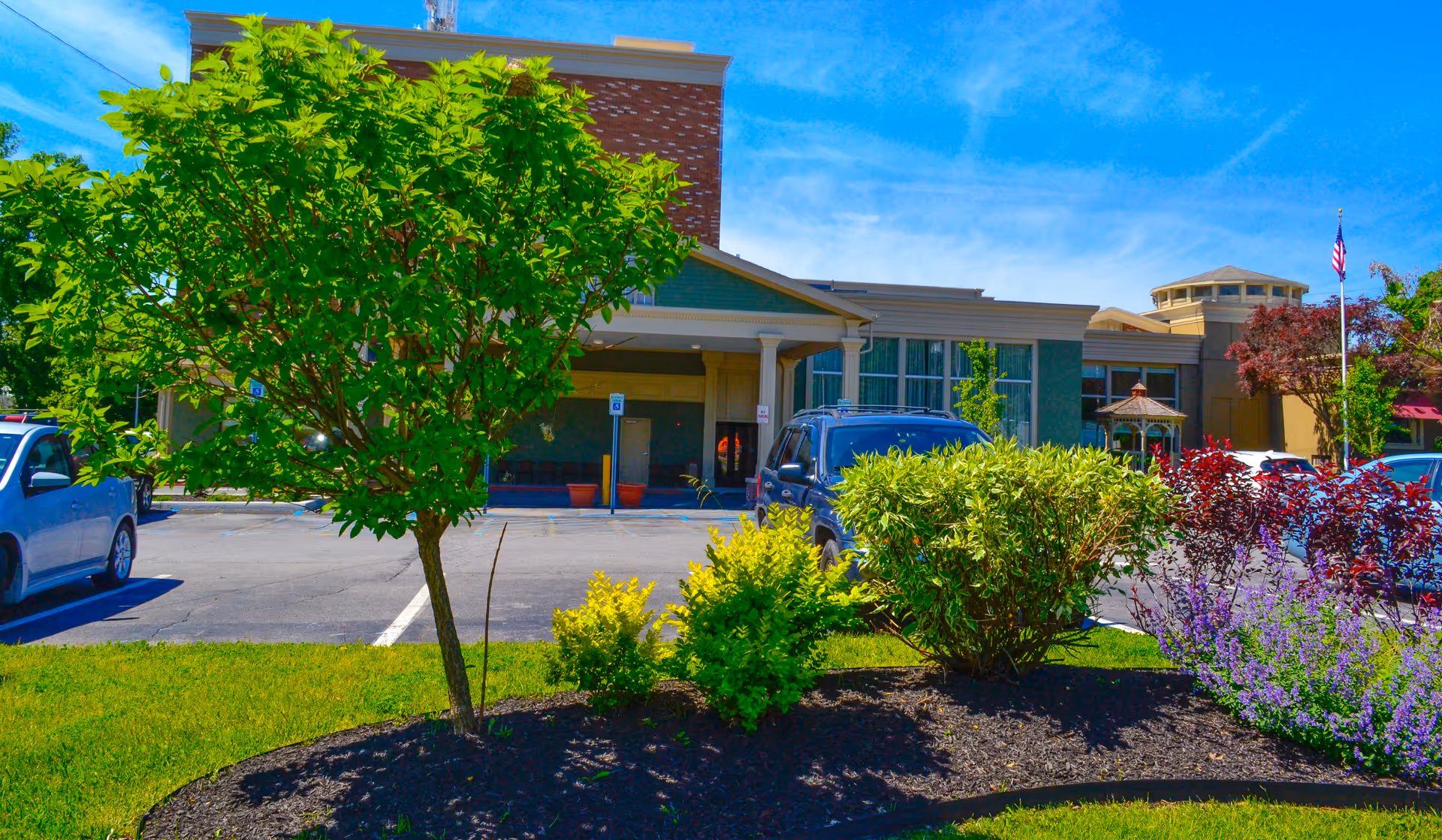Exterior view of Promenade at University Place facility showing the front entrance with a parking lot, several cars, a small landscaped area with green bushes and a tree, and a clear blue sky.